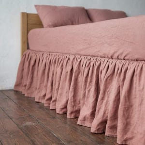 Peut inclure: Un cache-sommier rose poudré avec un design froncé et volanté qui pend de la base d'un cadre de lit. Le tissu présente un tissage texturé visible et recouvre la partie inférieure du lit, descendant jusqu'au sol en plis verticaux souples.