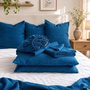 May include: A set of deep blue bedding including three square throw pillows, a fitted sheet, a flat sheet, and two rectangular pillowcases. The fabric has a visible woven texture, and the items are arranged neatly on a white bedspread.