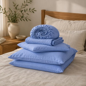 Peut inclure: Un ensemble de linge de lit bleu pervenche empilé sur un lit, comprenant un drap-housse roulé, un drap plat plié et deux taies d'oreiller rectangulaires. Le tissu présente une texture tissée visible et les articles sont disposés en pile.