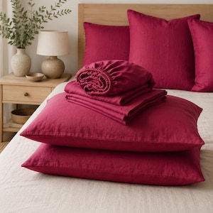May include: A set of deep magenta bedding including two stacked rectangular pillowcases, a folded flat sheet, and a fitted sheet with elastic edges. The bedding is displayed on a light beige textured bedspread with matching magenta pillows.