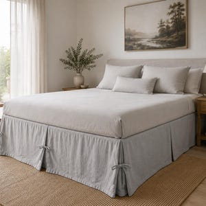Può includere: Una gonna per letto grigio chiaro caratterizzata da un design plissettato con chiusure decorative a fiocco su ogni angolo. Il tessuto ha una consistenza morbida e opaca e scende dalla base del materasso fino al pavimento, rifinendo il giroletto.