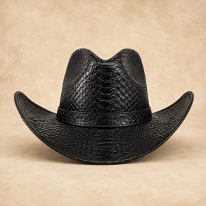 Puede incluir: Un sombrero de vaquero negro con un patrón texturizado de piel de serpiente en toda su superficie. El sombrero tiene una copa estructurada con un pliegue central y un ala ancha y curva. Una banda negra a juego rodea la base de la copa.