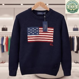 May include: A navy blue long-sleeve knit sweater featuring a crew neckline and a ribbed hem and cuffs. The center of the chest displays a large, knitted American flag graphic in red, white, and blue, with the red letters RL embroidered below it.