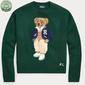 May include: A dark green long-sleeve crewneck sweater featuring a central graphic of a teddy bear wearing a navy blue varsity jacket, a patterned sweater, and cream-colored trousers. The letters RL are embroidered in white at the bottom right.
