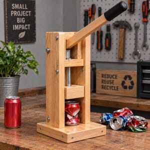 May include: A wooden can crusher featuring a vertical frame, a base, and a long lever handle with a black grip. The device is positioned on a wooden surface with a red aluminium can inside the crushing mechanism and several crushed cans nearby.