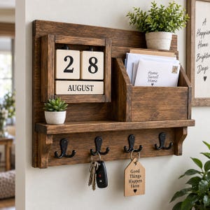 May include: A wall-mounted dark wood organizer featuring a perpetual block calendar displaying August 28, a mail slot containing envelopes, a small shelf with a potted succulent, and four black metal hooks holding keys and a wooden tag reading Good Things Happen Here.