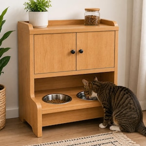 May include: A light oak-colored wooden pet feeding station featuring a cabinet with two doors and two black knobs. The lower section contains two circular stainless steel bowls embedded in the shelf, with a tabby cat eating from one bowl.