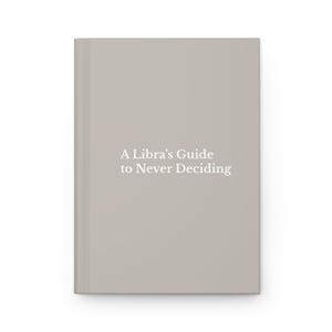 以下が含まれることがあります： 表紙の中央に「A Libra's Guide to Never Deciding」という白い文字が印刷された、ライトグレーのハードカバーノート。ノートは無地のシンプルな表面で、左側には縦の背表紙が見えています。