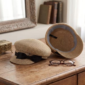 May include: Two straw newsboy caps with a natural tan color. One cap is positioned upright, featuring a black ribbon bow and a small metallic stud on the front. The second cap is tilted, revealing its interior with a grey lining and black strap.
