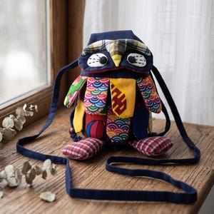May include: A handcrafted fabric bag shaped like an owl, featuring a dark blue top flap with a plaid accent, black button eyes, and a body covered in colorful, patterned fabric patches. It includes a long, thin dark blue shoulder strap.