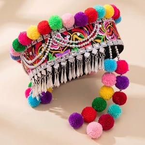 May include: A cylindrical headpiece featuring a black fabric base with vibrant floral embroidery. The top edge is lined with multicolored pom-poms. Silver-toned metal chains and dangling geometric charms adorn the front, with two long strands of pom-poms hanging from the side.