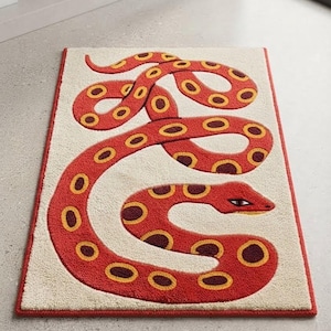 Puede incluir: Una alfombra rectangular con fondo color crema y borde rojo. La alfombra muestra un gráfico de una serpiente grande y enroscada en tonos rojos, amarillos y marrón oscuro, con un detalle distintivo de la cabeza y el ojo en el lado derecho.