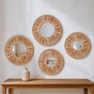 May include: A set of four circular wall mirrors featuring light brown, radiating rattan or reed frames. Each mirror has a central glass pane surrounded by thin, closely spaced spokes that create a sunburst effect. The mirrors vary in size.