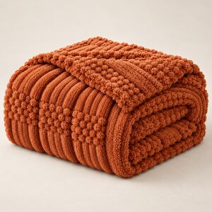 May include: A folded burnt orange throw blanket featuring a textured pattern of alternating vertical ribbed stripes and square sections of raised bobble stitches. The fabric has a soft, plush appearance with a consistent deep orange color throughout.