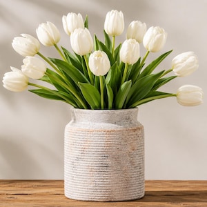 May include: A bouquet of white tulips with green stems and leaves arranged in a cylindrical, textured, light gray stone vase. The vase features horizontal ribbed detailing and a weathered, rustic finish, holding the flowers upright in a display.