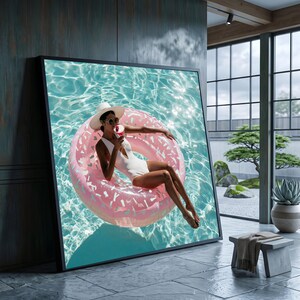 Op de afbeelding: Een grote ingelijste kunstprint aan de muur met een persoon in een wit badpak, een hoed met brede rand en een zonnebril, ontspannend op een roze donutvormige zwemband in turquoise water terwijl diegene een roze drankje vasthoudt.