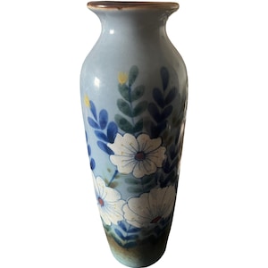 May include: A light blue ceramic vase featuring a hand-painted floral pattern with white flowers, dark blue leaves, and small yellow accents. The vase has a flared rim with a brown edge and a tapered base that transitions into a darker brown tone.