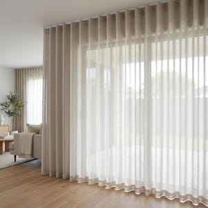 May include: Floor-to-ceiling beige sheer curtains with a vertical wave fold design. The curtains are hung from a ceiling-mounted track, creating uniform pleats that extend from the top to the floor, allowing soft light to filter through the fabric.