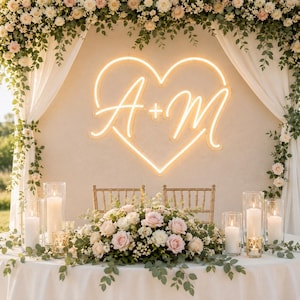 May include: A glowing warm white neon sign in the shape of a heart with the letters A + M in the center. The sign is mounted on a wall behind a table decorated with a large floral arrangement of pink and white roses and several glass candles.