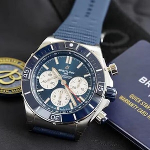 May include: A Breitling Chronomat watch with a blue dial, three white sub-dials, and a blue rubber strap. The watch features a rotating bezel with a tachymeter scale, a date window, and luminous hands. A Breitling warranty card holder is included.