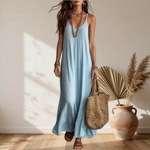 Puede incluir: Un vestido largo de color azul claro con tirantes finos y una silueta holgada y fluida. El vestido se combina con un bolso de paja trenzada, collares dorados superpuestos y sandalias marrones, creando un conjunto sencillo y coherente.