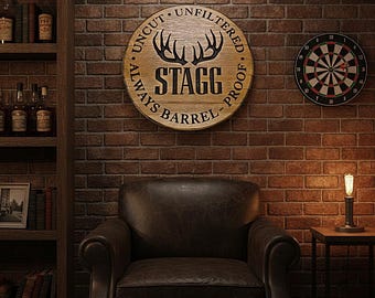 May include: A circular wooden wall sign featuring the text STAGG, an illustration of deer antlers, and the words UNCUT, UNFILTERED, ALWAYS BARREL PROOF. The sign is mounted on a dark brown brick wall above a leather armchair in a dimly lit room.