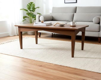 May include: A rectangular wooden coffee table with four tapered legs and a smooth, medium-brown finish. The table surface holds a stack of books, a small white bowl containing wooden beads, and a single white ceramic mug on a small saucer.