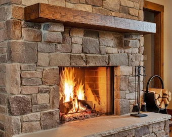 May include: A stone fireplace featuring a large, dark brown wooden mantelpiece above a burning fire. The hearth is constructed from flat, light-colored stone blocks. A black metal fireplace tool set and a circular log holder filled with wood sit nearby.