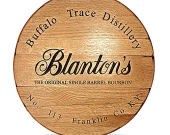 May include: A circular wooden barrel head featuring the text Buffalo Trace Distillery and Blanton's The Original Single Barrel Bourbon. The wood has a natural grain pattern with the text No. 113 and Franklin Co KY engraved in black lettering.