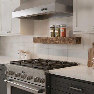 May include: A rustic, dark brown wooden floating shelf mounted on a white tiled kitchen wall above a stainless steel gas range. Three glass spice jars with silver lids sit on the shelf, containing green herbs and red spices, under a range hood.