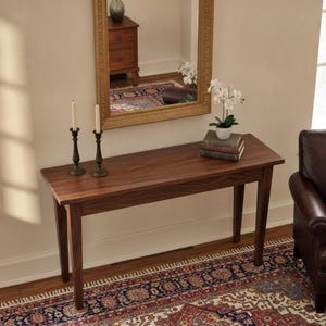 May include: A rectangular wooden console table with four straight legs, featuring a dark brown finish. Two tall, slender metal candle holders stand on the left side, while a stack of three books and a small white potted orchid sit on the right.