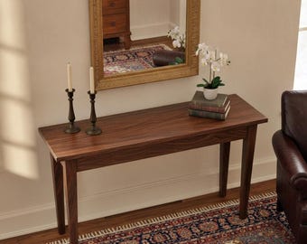 May include: A rectangular dark brown wooden console table with four straight legs. On the table surface are two tall dark metal candle holders with white candles, a stack of three books, and a small white pot containing a white orchid plant.