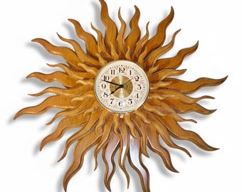 May include: A sunburst wall clock featuring a circular clock face with black Arabic numerals and ornate gold-toned center detailing. The clock is surrounded by multiple layers of carved wooden rays in a natural light brown finish.