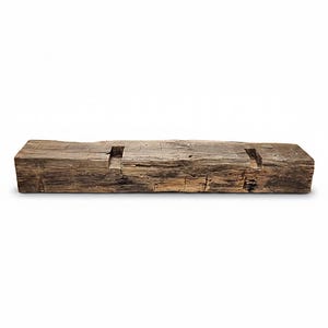 May include: A rectangular piece of reclaimed wood with a weathered, dark brown finish and a rough, textured surface. The top side features two evenly spaced, rectangular cutouts carved into the wood, showing natural grain and deep cracks.