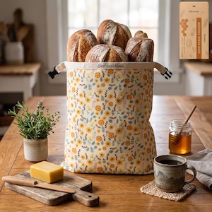 Könnte beinhalten: Ein wiederverwendbarer Bienenwachs-Brotbeutel mit cremefarbenem Hintergrund und gelb-grünem Blumenmuster. Der Beutel trägt den Schriftzug BeeFreshHome am oberen Rand und hat schwarze seitliche Schnallen. Er ist mit vier Brotlaiben gefüllt.