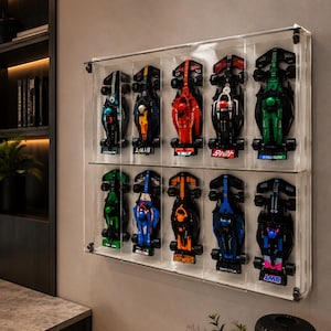 May include: A rectangular clear acrylic wall-mounted display case containing ten miniature Formula 1 race cars arranged in two horizontal rows. Each car features distinct colour schemes including red, green, blue, black, and orange, with visible driver cockpits.