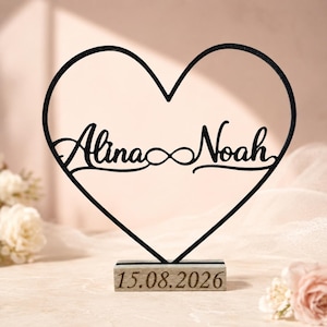 Könnte beinhalten: Ein schwarzer herzförmiger Drahtrahmen mit den Namen Alina und Noah in Schreibschrift und einem Unendlichkeitssymbol dazwischen. Der Rahmen steht auf einem rechteckigen Holzsockel, in dessen Vorderseite das Datum 15.08.2026 eingraviert ist.