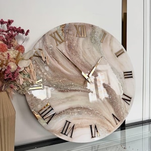 Può includere: Un orologio da parete rotondo con una superficie lucida e marmorizzata nei toni del crema, beige e rosa tenue, con numeri romani dorati. Il quadrante presenta una finitura testurizzata simile alla resina con accenti metallici e lancette sottili.