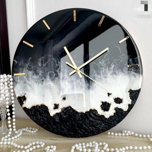 Könnte beinhalten: Eine runde Wanduhr mit glänzender schwarz-weißer Harzoberfläche in Marmoroptik. Das Zifferblatt hat goldfarbene Stundenmarkierungen und passende goldene Zeiger. Der untere Bereich weist eine strukturierte, mattschwarze Oberfläche auf.