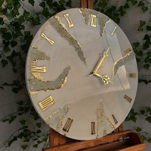 May include: A circular wall clock with a smooth beige face featuring gold Roman numerals and decorative fissures filled with clear, textured crystalline accents. The clock has gold-colored hour, minute, and second hands centered on the face.