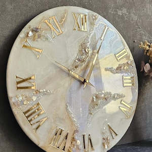 May include: A round wall clock featuring a cream and white marbled resin face with gold-colored Roman numerals. The clock face is accented with embedded crushed stone and shell fragments, and it includes thin, pointed gold-colored metal clock hands.