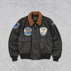 Puede incluir: Una chaqueta tipo bomber de cuero marrón oscuro con cuello de piel sintética marrón, cierre de cremallera frontal y dos grandes bolsillos con solapa. La chaqueta está decorada con varios parches bordados de estilo militar en el pecho y las mangas.