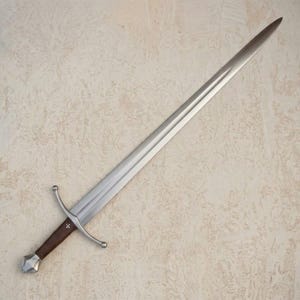 Op de afbeelding: Een stalen zwaard met een lang, tweesnijdend lemmet met een centrale groef. Het gevest heeft een pareerstang met gebogen stootplaten, een bruine, met leer omwikkelde greep en een metalen pommel. Een klein wit kruis is zichtbaar op de greep.