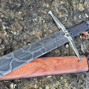 Puede incluir: Una espada con una hoja oscura estampada que presenta un diseño gris similar a una red, una guarda plateada y un mango envuelto en negro. La espada está colocada junto a una vaina de madera lisa de color marrón rojizo con un cordón negro.