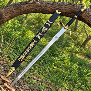 Puede incluir: Una espada de metal plateado con mango envuelto en negro y guarda dorada con extensiones en forma de alas. La vaina negra a juego muestra intrincados símbolos y patrones dorados, incluido un emblema de trifuerza central.