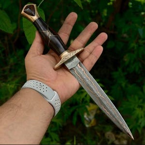 May include: A double-edged dagger featuring a dark wood handle with gold-coloured metal accents and a patterned Damascus steel blade. The blade displays a distinct wavy texture with a central copper-coloured inlay running down the length of the steel.