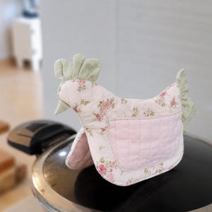 May include: A quilted fabric chicken-shaped tea cozy featuring a light pink background with a floral rose pattern, a solid light pink pocket on the side, and light green fabric accents for the comb, beak, and tail feathers.