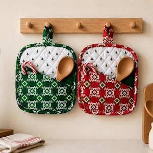 May include: Two square quilted pot holders with hanging loops, one in green and one in red, featuring a white diamond pattern with small green trees. Each has a front pocket containing a wooden spoon and a candy cane, hanging on a wooden rack.