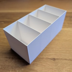 Könnte beinhalten: Ein rechteckiger Schreibtisch-Organizer aus weißem Kunststoff mit vier gleich großen, vertikalen Fächern. Der Behälter hat klare, gerade Kanten und ein offenes Design an der Oberseite, um verschiedene kleine Gegenstände zu sortieren.