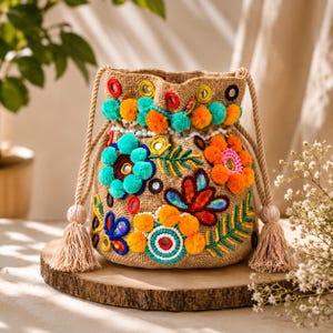 May include: A tan hessian drawstring bag featuring vibrant embroidery, colourful pom-poms, and small circular mirrors. The bag is adorned with floral patterns in orange, turquoise, and red, finished with thick rope handles and matching tan tassels on each side.